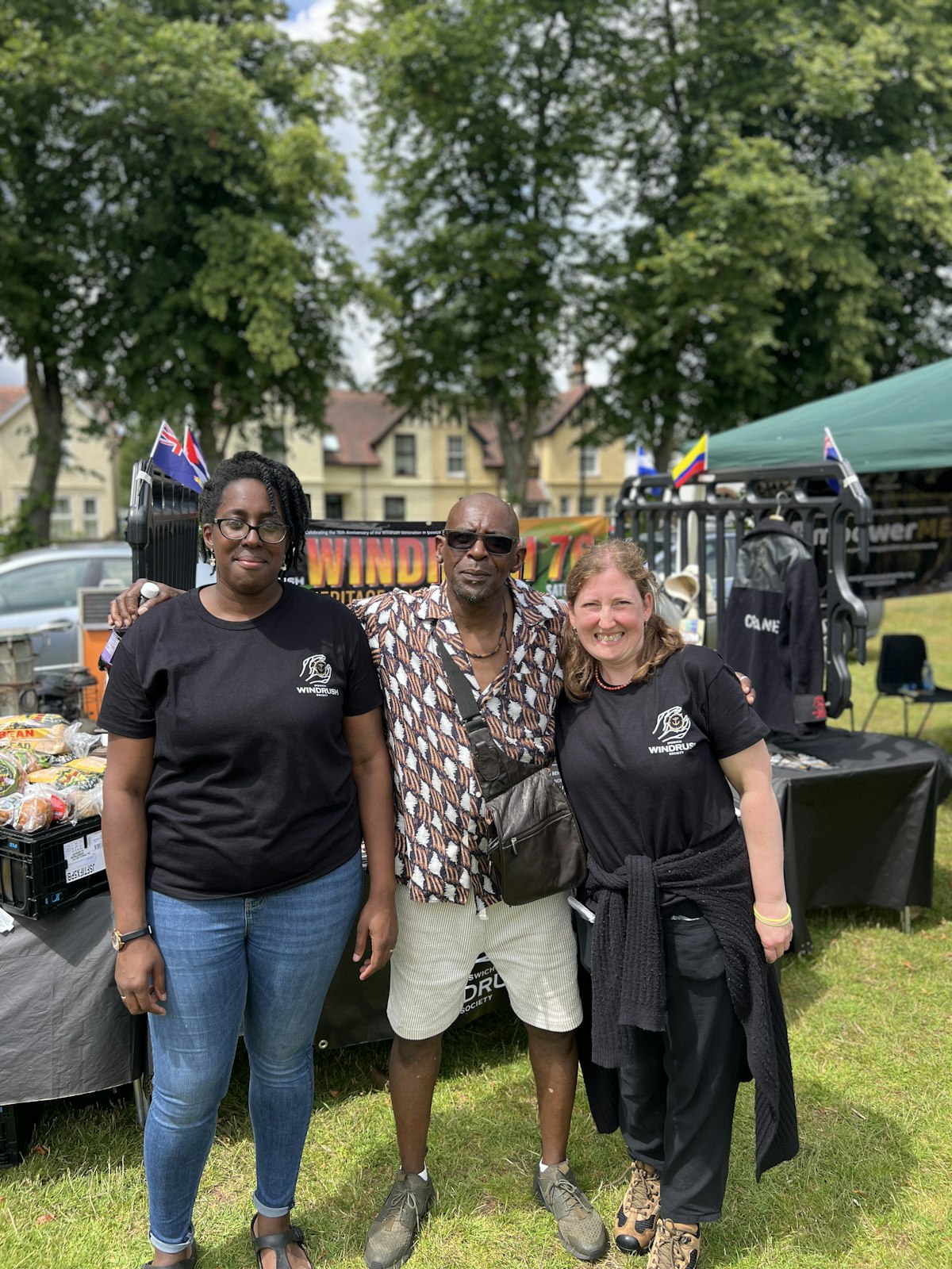Three Windrush festival organisers pose together wearing event t-shirts with festival banners and stage visible in background